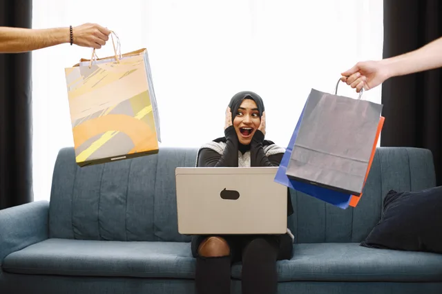 A woman in a hijab excitedly shopping online, receiving bags while sitting on a couch with a laptop. How ecommerce SEO business models differ without a set pricing rule